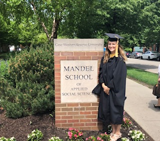 Rachel Truhan in graduation cap and gown standing in front of a sign for the Mandel School of Applied Social Sciences