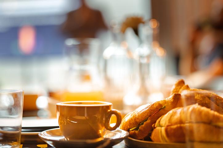 A tan cup of coffee next to a croissant with glassware in the background