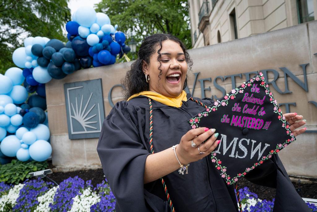 Woman holds her graduation cap in front of the CWRU sign on Euclid with balloons