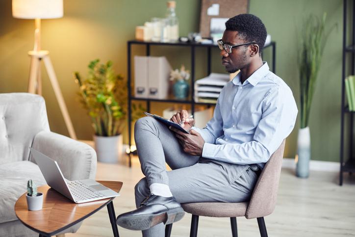 Person sits in a chair and takes notes while looking at a laptop on a table