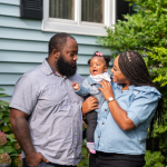 Two parents holding a baby outside a house