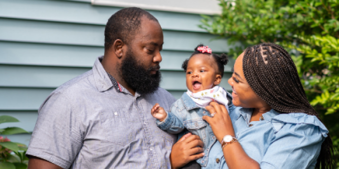 Two parents holding a baby outside a house