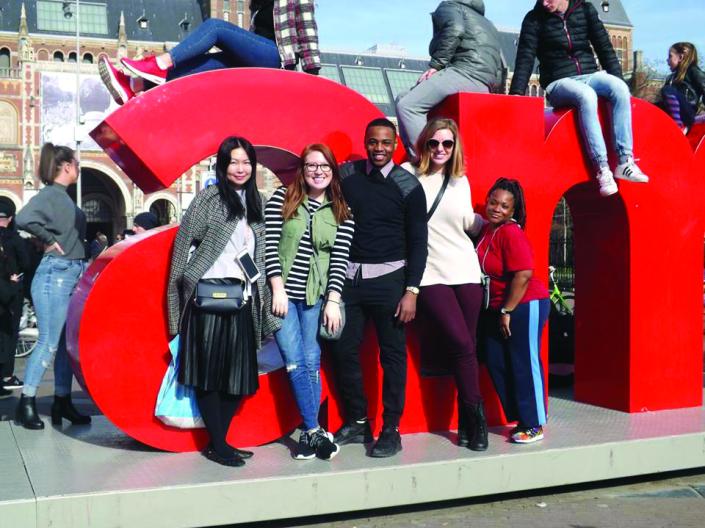 Image of five students standing in front of a large red lowercase letter A and large lowercase letter N, with three students sitting above