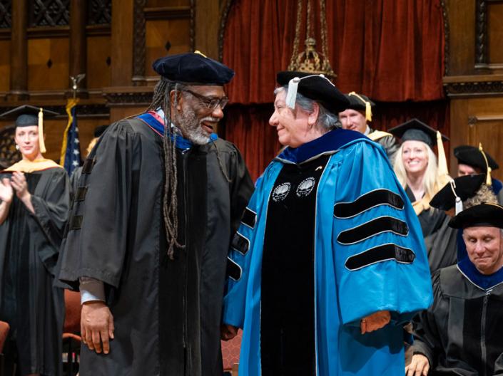 David Miller and Marji Edguer smile at each other and hold hands on stage at commencement 2025
