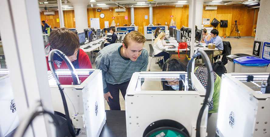 Three college students lean down to look inside 3D printing machines in front of students working at rows of computers.
