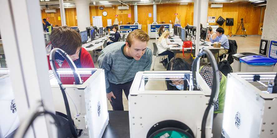 Three college students lean down to look inside 3D printing machines in front of students working at rows of computers.