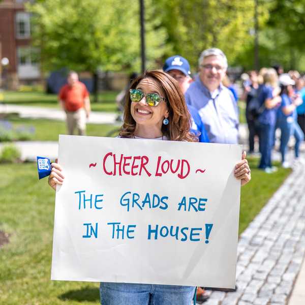 A woman holding a sign that reads, “Cheer loud, the grads are in the house!”
