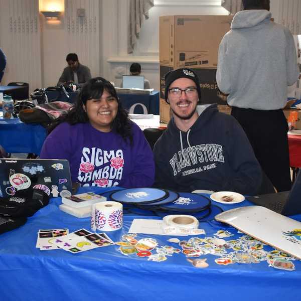 Two CWRU students pose for a photo seated at a table covered in stickers and other giveaway items.