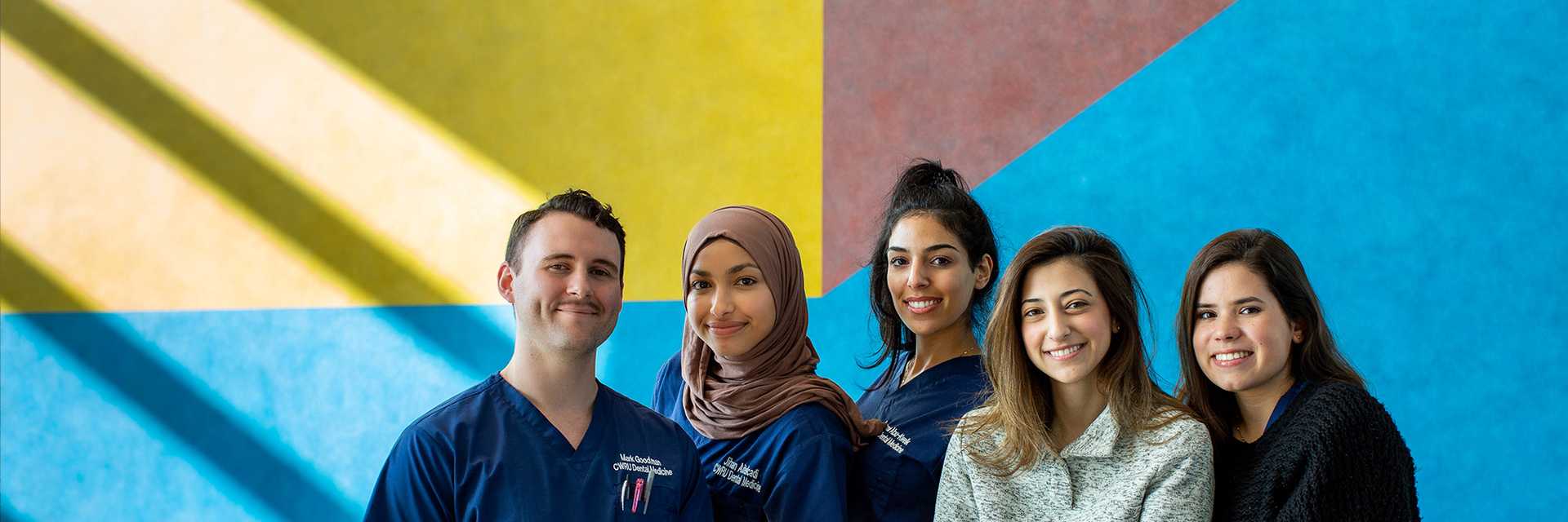 Photo of five Case Western Reserve University students wearing scrubs stand in front of a mural in the Health Education Campus