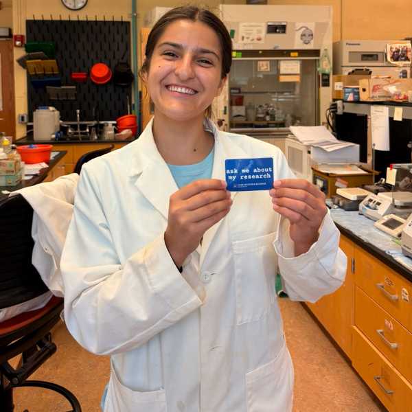 Fourth-year undergraduate student, Emilia Sanz-Rios, posing and holding a sticker that reads, “Ask me about my research.”