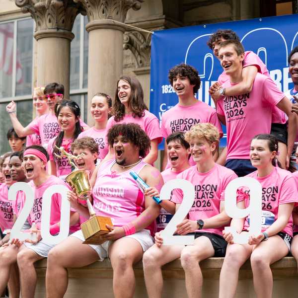 A group of first-year students wearing pink “Hudson Relays 2026” shirts pose outside Adelbert Hall, cheering and holding a trophy alongside white cutout numbers reading “2029.”