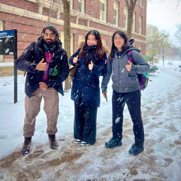 A group of three students, posing with their thumbs up, smiling as snow fell on campus.