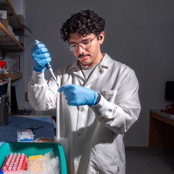 A student in a white lab coat and blue gloves uses a pipette to transfer liquid into a small tube while working in a laboratory.