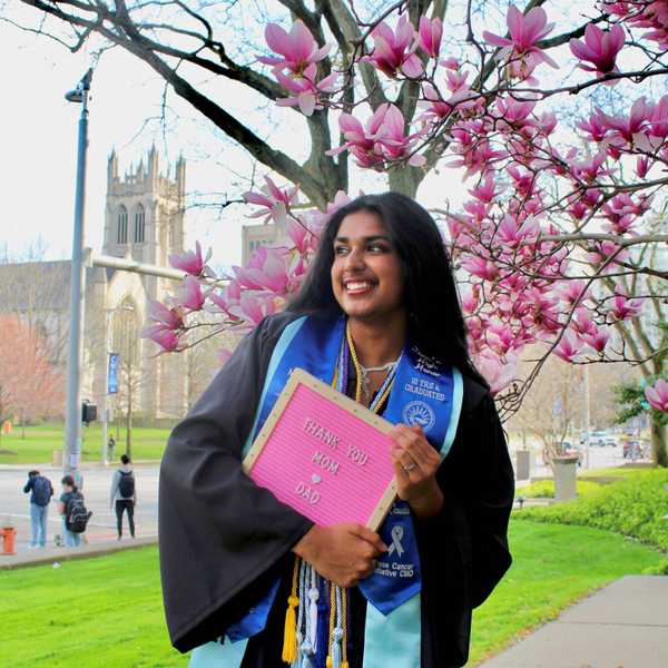 One woman student, Bhavya Bansal, poses in front of a magnolia tree wearing academic regalia with a pink sign that reads, “Thank you Mom and Dad.”