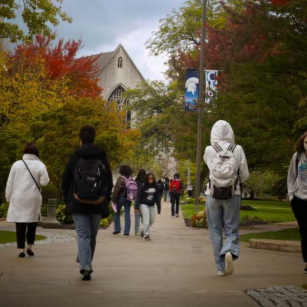 Numerous CWRU students walk to and from classes on Case Quad on a sidewalk surrounded by trees with leaves changing colors.