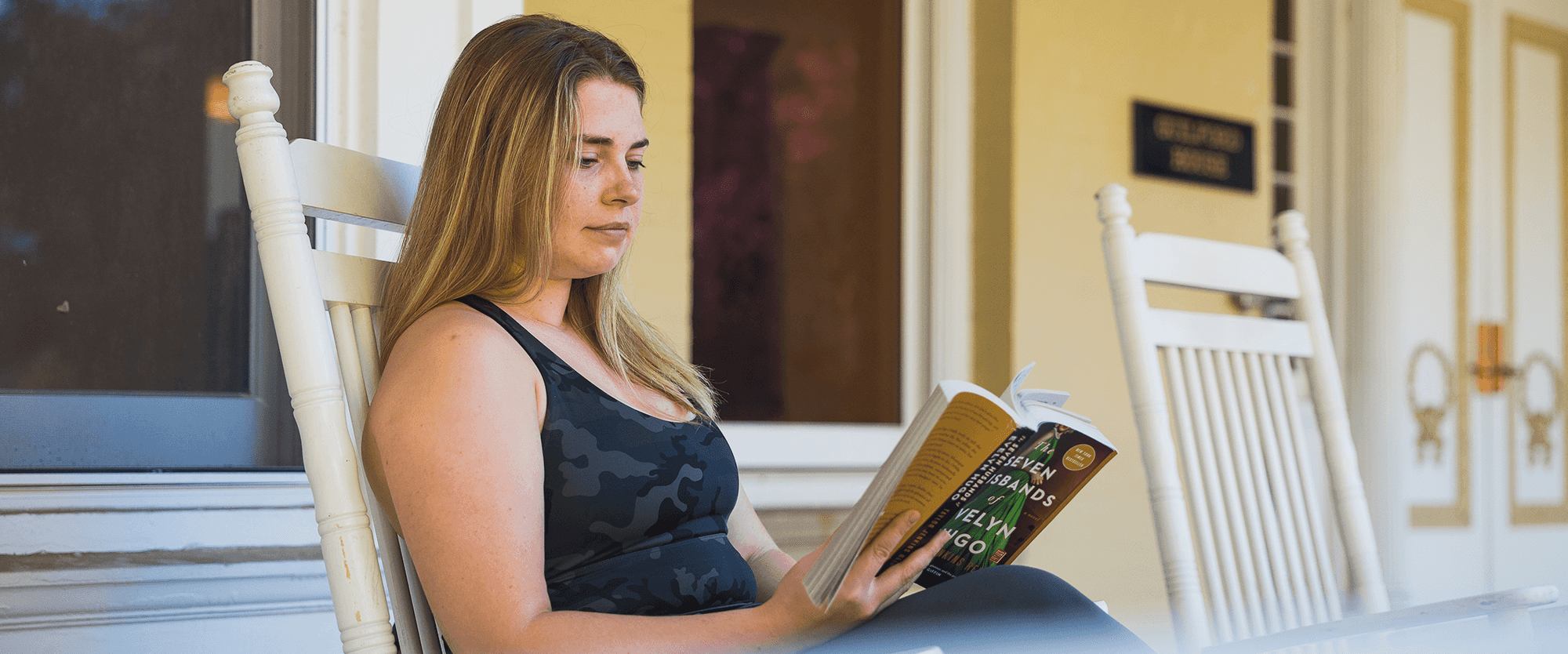 Woman reading a literature book outside on a patio while sitting in a rocking chair.