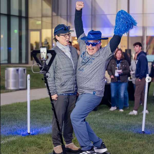 Two CWRU parents pose in a 360 photo booth with blue light up sunglasses, a blue cowboy hat and a pompom during Blue Bash. 