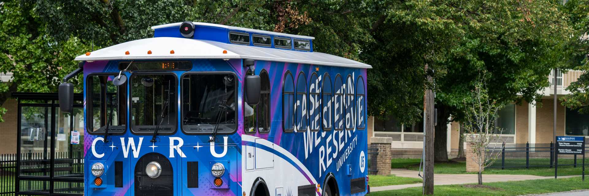 A blue CWRU trolley travels along a tree-lined campus road