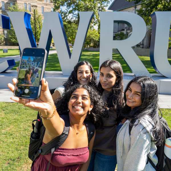 A group of 4 students pose for a selfie in front of a large gray and blue sign that spells, “CWRU.”