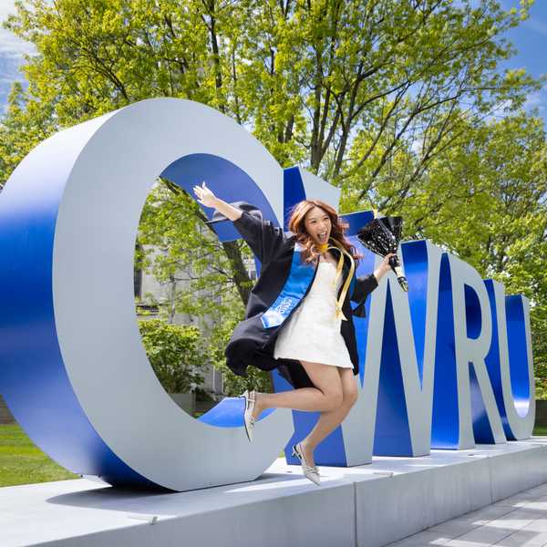 A student jumping in a cap and gown in front of the “CWRU” sign on campus.