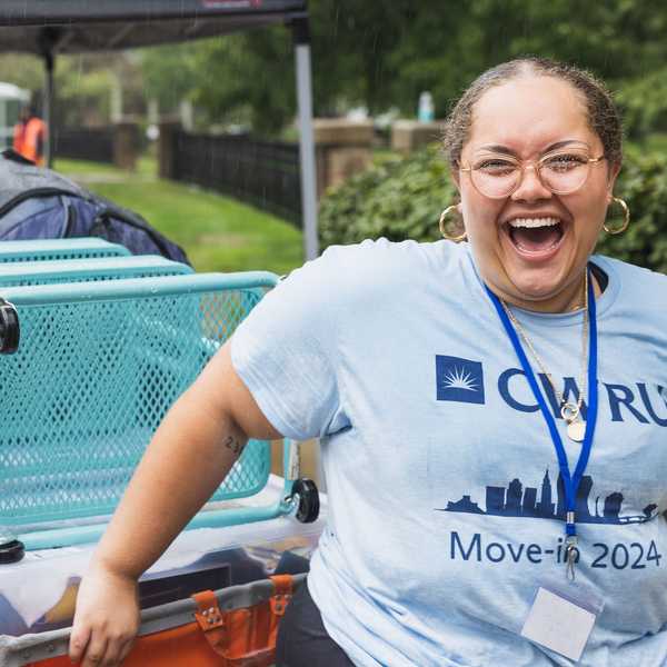 Two student volunteers smile in the rain while pushing a cart of dormitory supplies inside during move-in weekend.