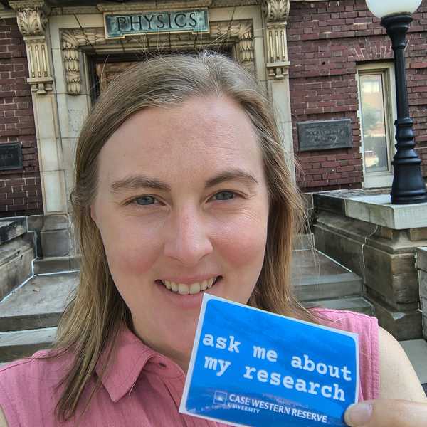 CWRU researcher Lydia Kisley posing for a selfie holding a sticker that reads, “ask me about my research.”