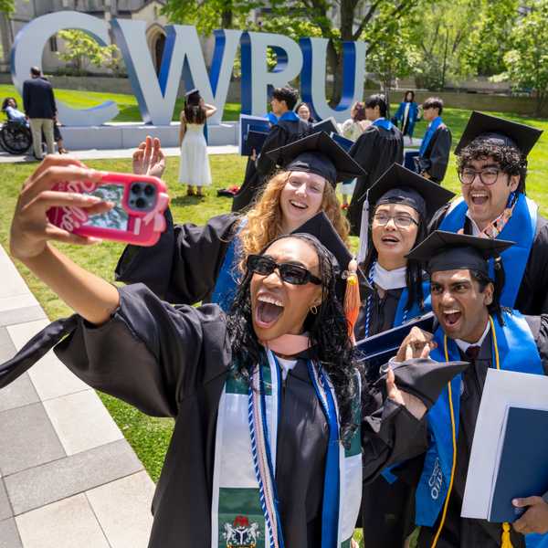 A group of five students in caps and gowns posing for a selfie in front of the “CWRU” sign on campus.
