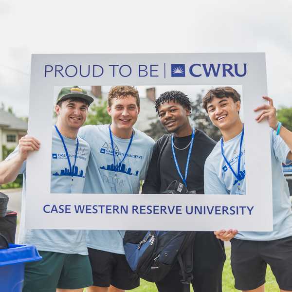 Four students holding a sign that reads, “Proud to be CWRU,” next to a cart of dormitory supplies during move-in weekend.