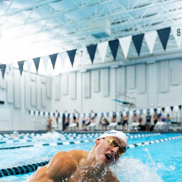 A swimmer wearing a swim cap and goggles splashes water on a turn in a pool set indoors beneath blue and white pennants. 