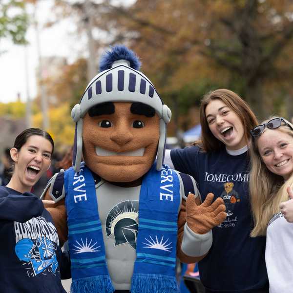 A group of three students smiling and posing with CWRU’s Spartie mascot during homecoming.