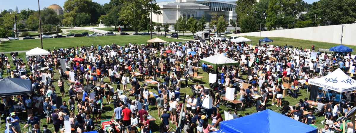 Large outdoor field on a sunny day filled with tables. Many students gathered listening and laughing with each other.