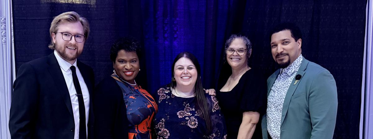 Five people - three women and two men - stand in a row, smiling for a group photo against a backdrop. 