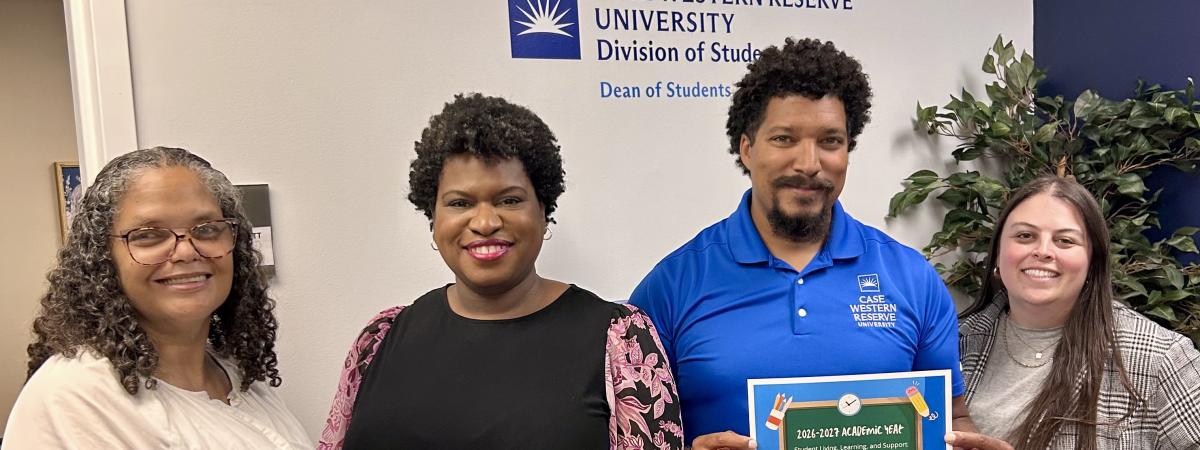 Four individuals standing together. One is in a white shirt, one is in a black shirt, one is in a blue polo, and one is in a striped blazer. They are holding a back to school sign under the dean of students office logo. 
