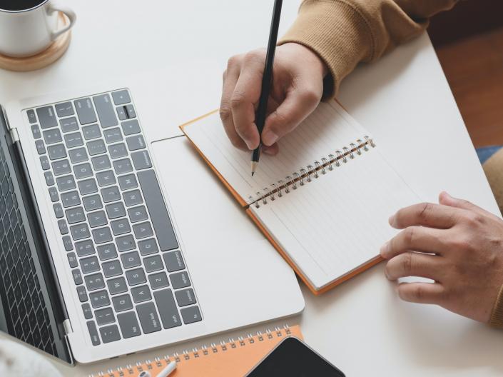 Close up of a desk with a computer and a person writing in a notebook