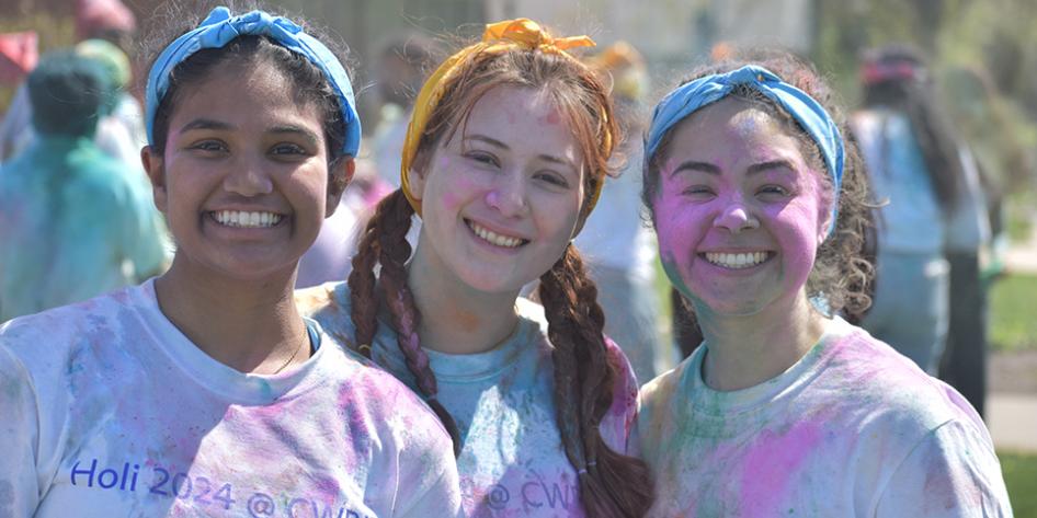 Three smiling students in white shirts covered in vibrant colored powder that reads Holi 2024 @ CWRU.