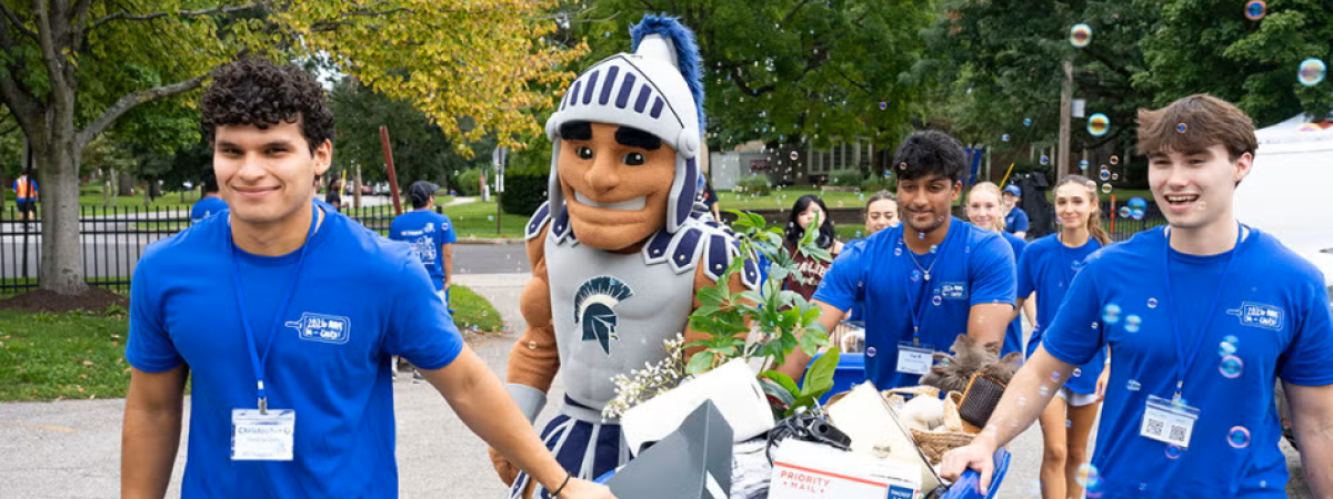 Student volunteers with Spartie pushing a moving cart during first year move-in day