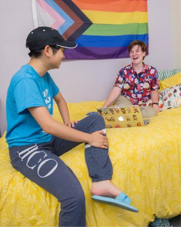 Two students sitting on a dorm bed in front of a flag with a rainbow of colors