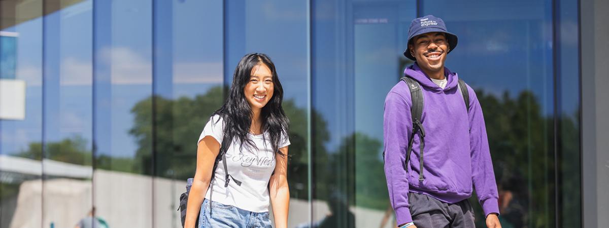 Two smiling students with backpacks walking in front of a modern glass building on a sunny day.