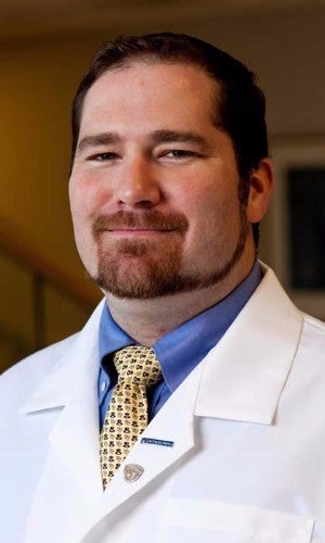 A middle-aged man with a beard and closed-lip smile wearing a lab coat and tie