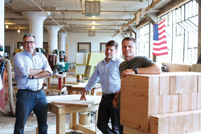 A photo of three men in a factory with wooden tables and blocks of wood