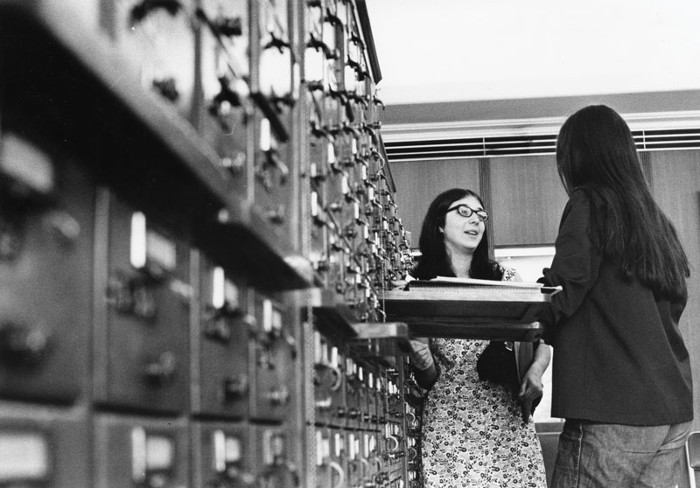 Black and white photo of two women standing next to a large filing cabinet with a drawer open