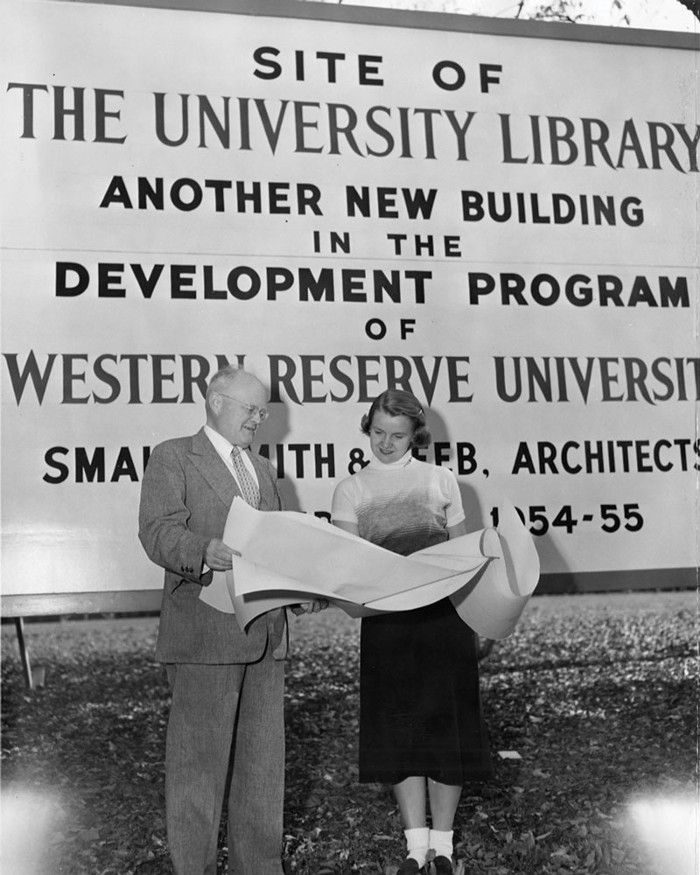 A black and white photo of a man and a woman reading from a long piece of paper in front of a new library billboard.