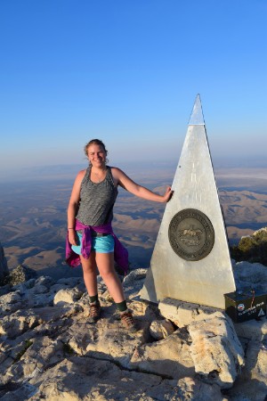 A female student standing at the top of Guadalupe Peak with her hand resting against a monument