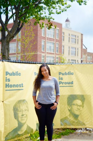 Female student standing with her hands on her hips in front of a fence with large signs that say: Public is Honest and Public is Proud