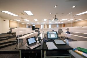 Picture of lectern devices and laptop set up in a classroom, behind acrylic glass