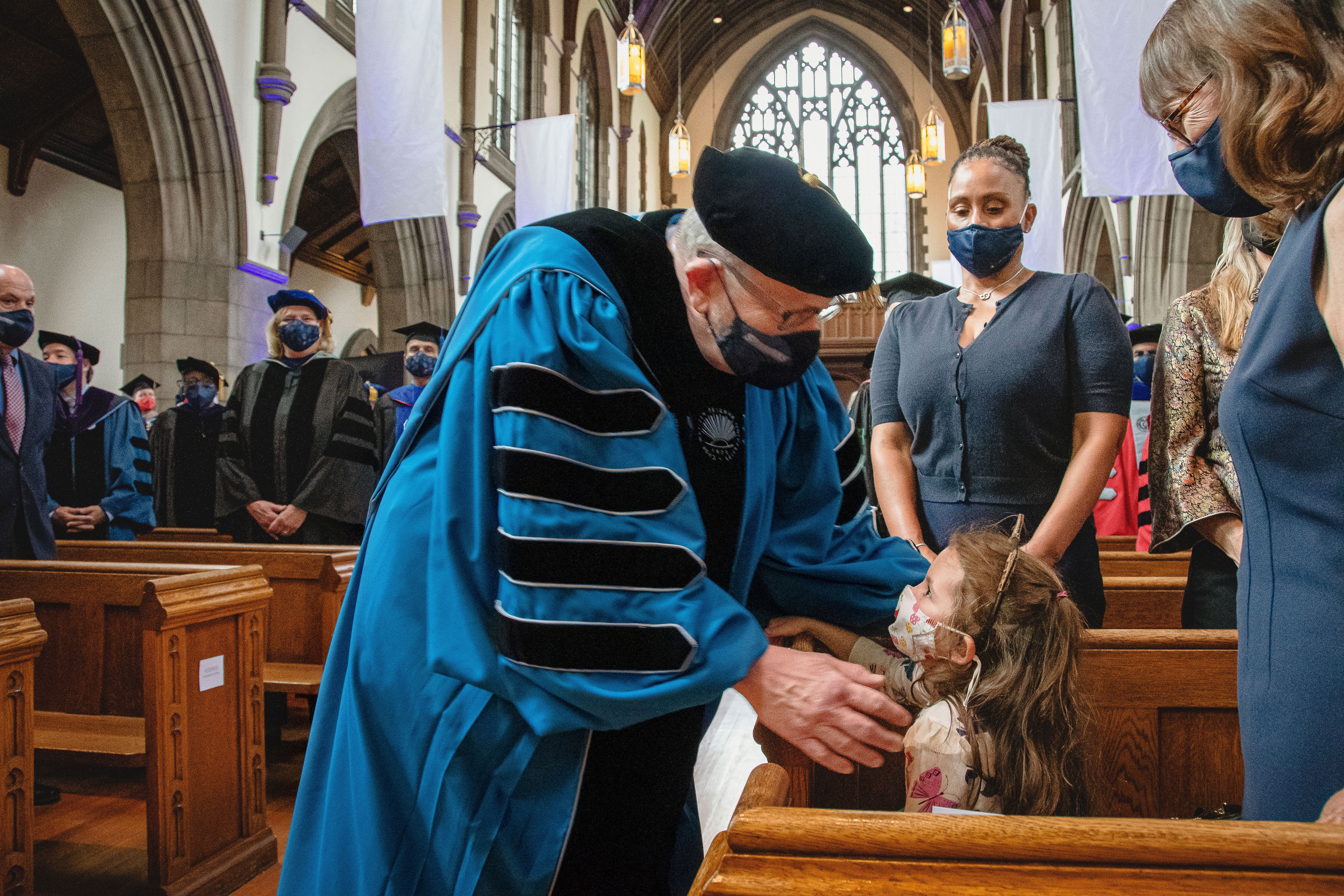 Eric Kaler, wearing a mask and commencement robes, hugs his granddaughter in a chapel before
      reaching the inauguration stage