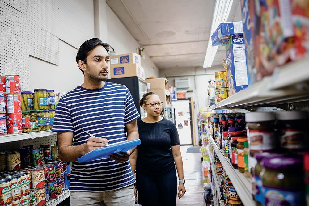 Two students walking in a store with a clipboard reviewing groceries on the shelves.