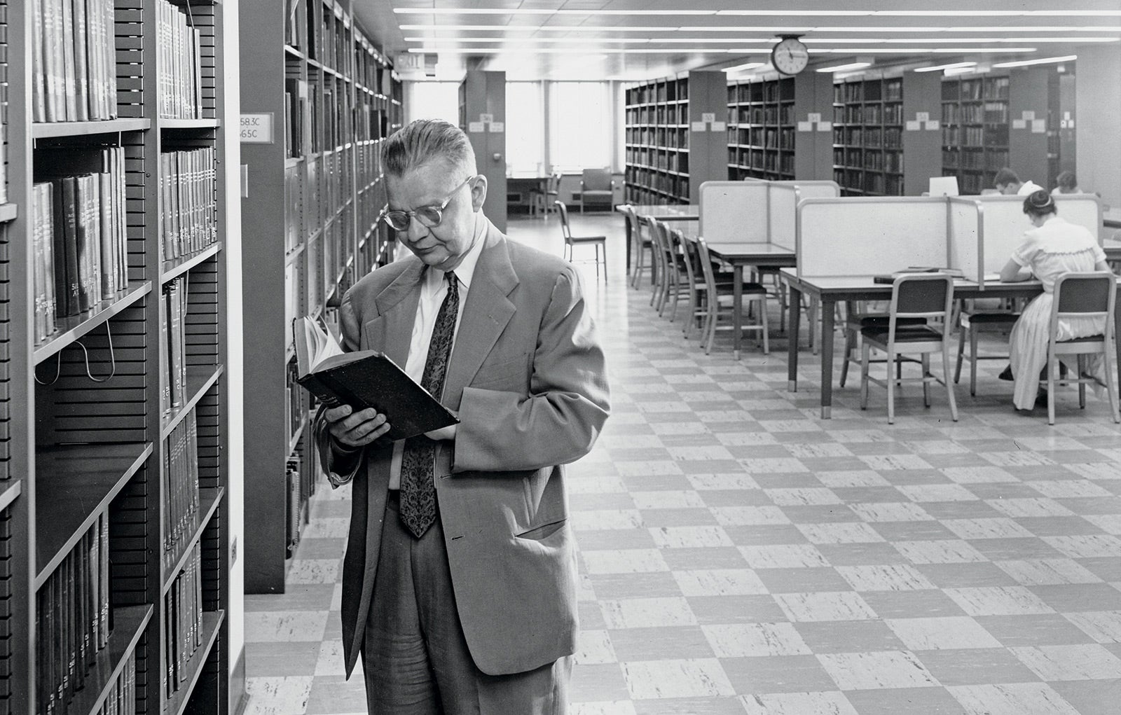 Black and white full body photo of Jesse Shera reading in the library.