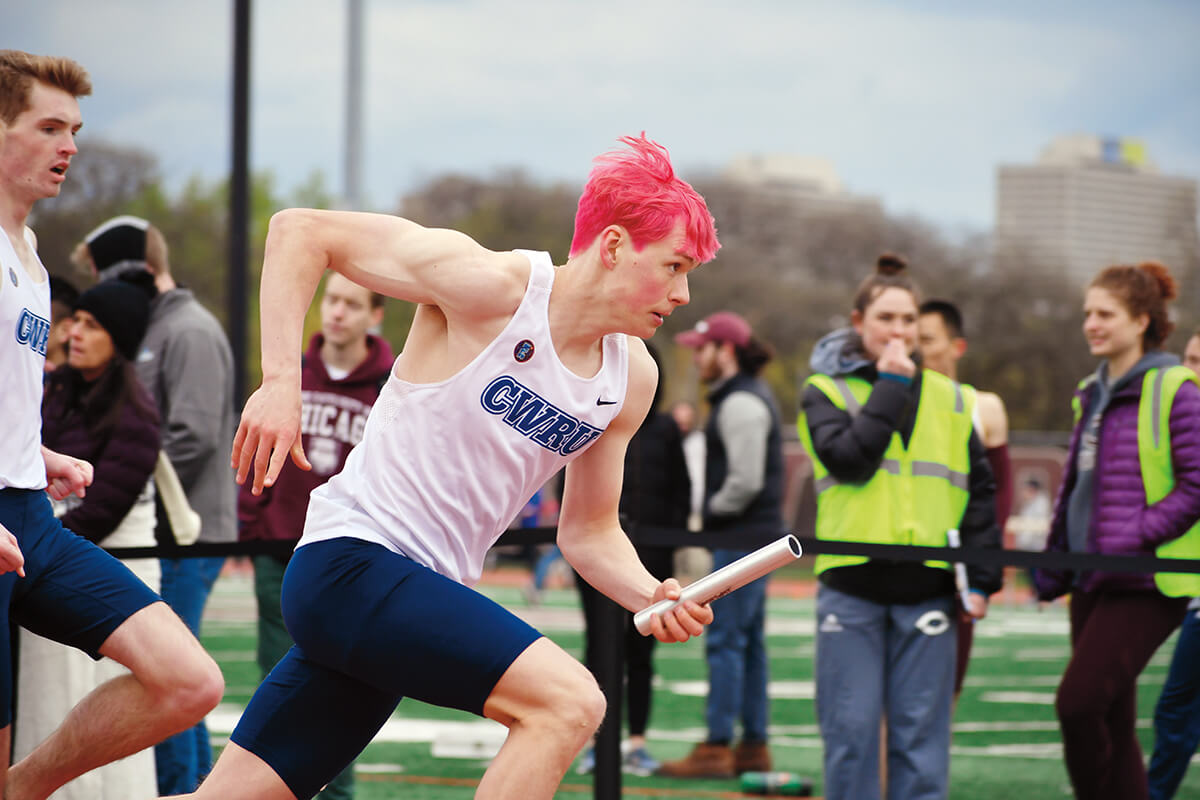 Photo of a student athlete running outdoors with a baton. People watch in the background.