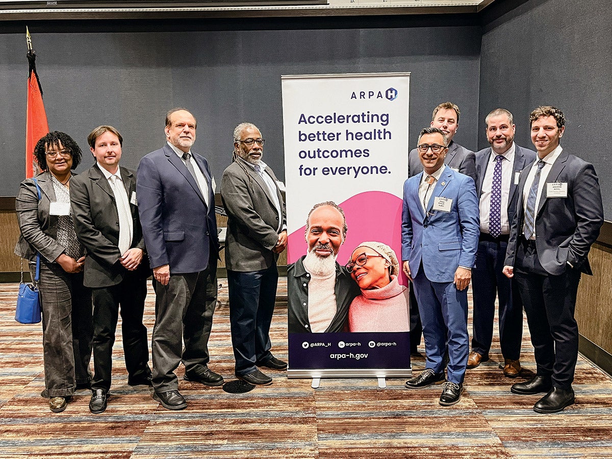 Eight people in a meeting room, four to the left and four to the right of a tall banner that reads: 'Accelerating better health outcomes for everyone.''